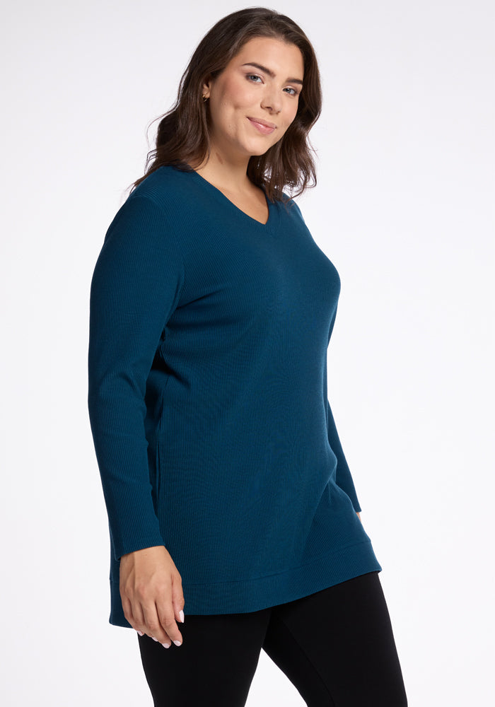 A person with long brown hair is wearing a Real Teal Evie Tunic from Woolx, known for its temperature-regulating Merino wool, paired with black pants. They are smiling and standing against a plain white background. 