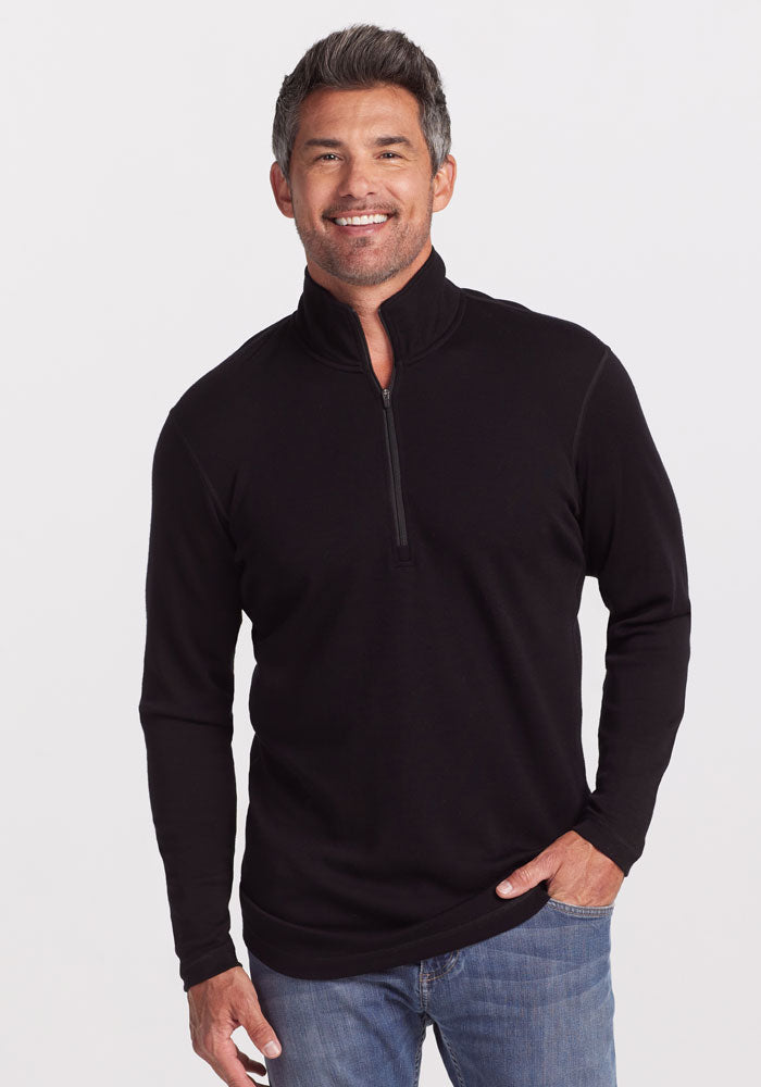 A person with short, gray hair is smiling and standing against a plain white background. They are wearing an ultra-soft Explorer 1/4 Zip pullover in black from Woolx, made of midweight fabric, paired with blue jeans. One hand is in their pocket while the other rests at their side.