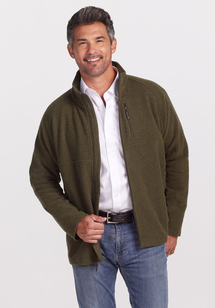 A man wearing a white shirt and an olive green Fairbanks Jacket by Woolx stands smiling with one hand in his jeans pocket, set against a plain gray background. 