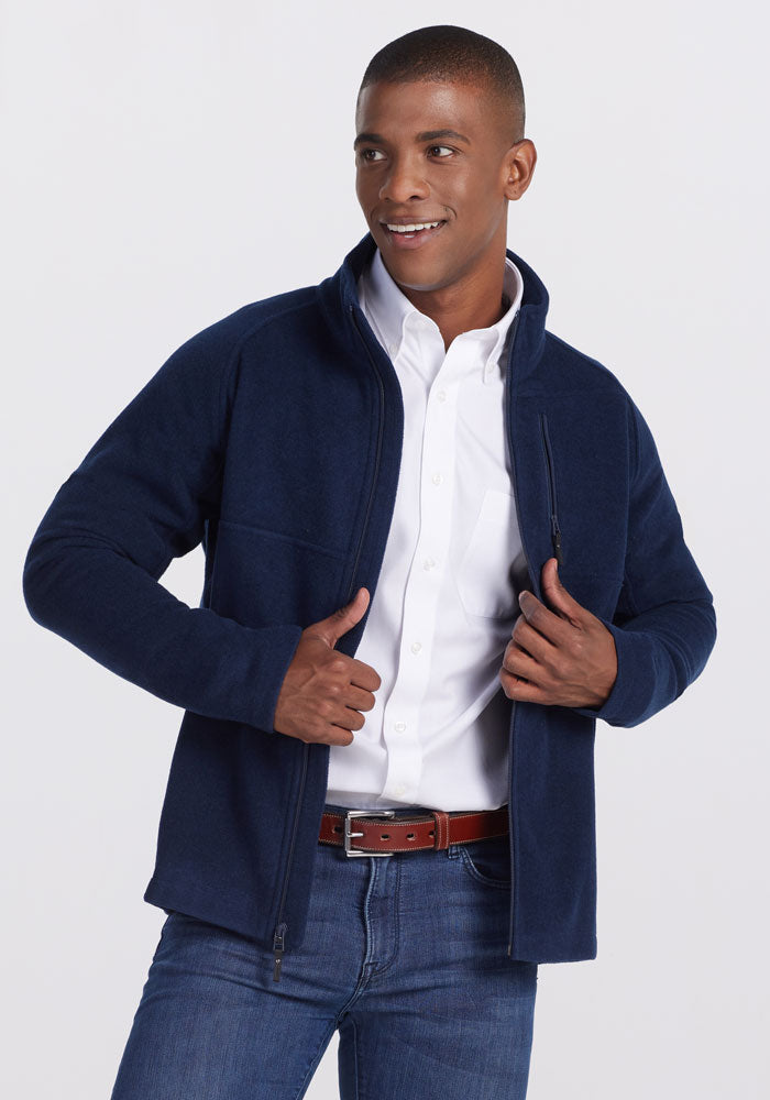 A person wearing a dark navy Fairbanks Jacket from Woolx over a white button-up shirt and blue jeans poses confidently. They are smiling and adjusting their ultra-heavyweight jacket with both hands against a plain, light-colored background. 