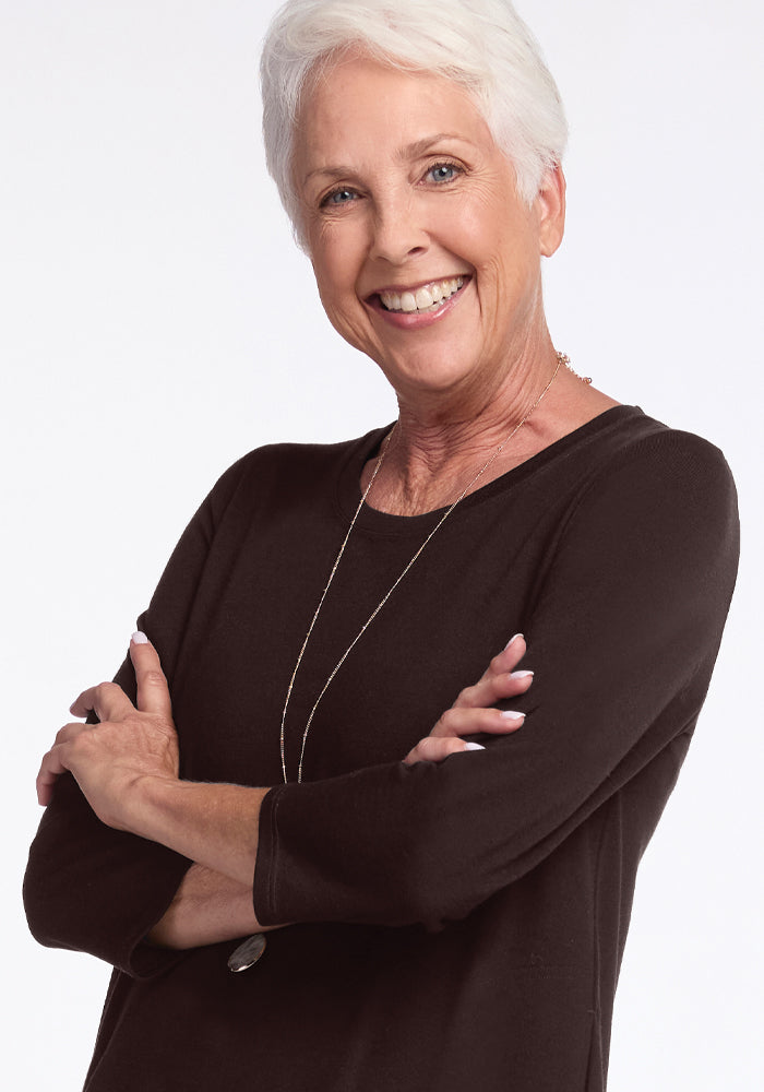 A smiling older woman with short white hair, adorned in a black top and a statement necklace, exudes confidence akin to the classic elegance of the Rissa A Line Dress by Woolx, her arms confidently crossed against a plain white background.