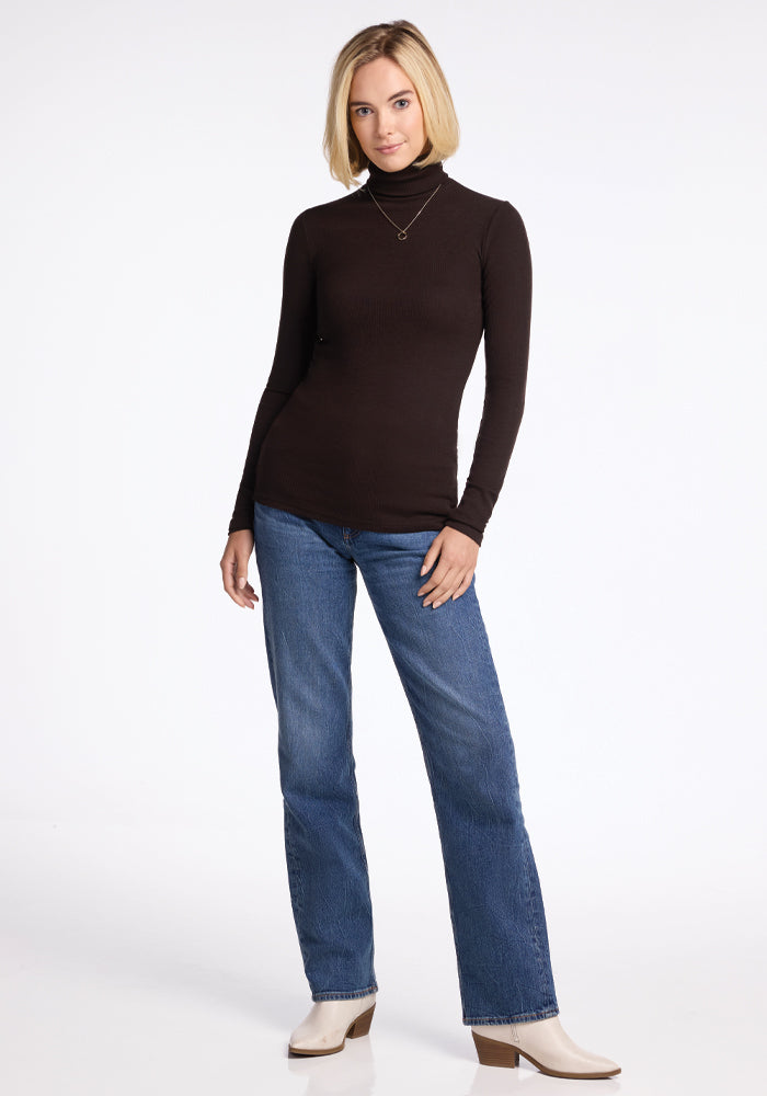 A person dressed in a Woolx Sage Ribbed Turtleneck in French Roast, paired with blue jeans, white boots, and a necklace stands against a plain white background. They have short, light-colored hair and are looking at the camera with a relaxed posture.