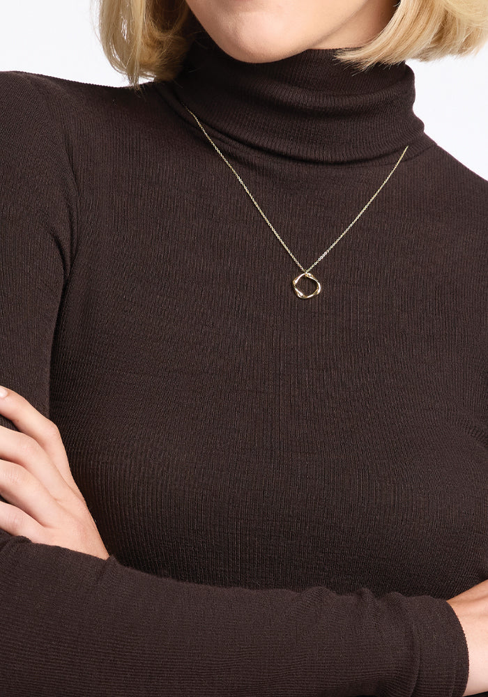 A person with blonde hair wearing a Sage Ribbed Turtleneck in French Roast by Woolx and a simple gold necklace. The arms are crossed, highlighting the ribbed fabric, against a plain white background.