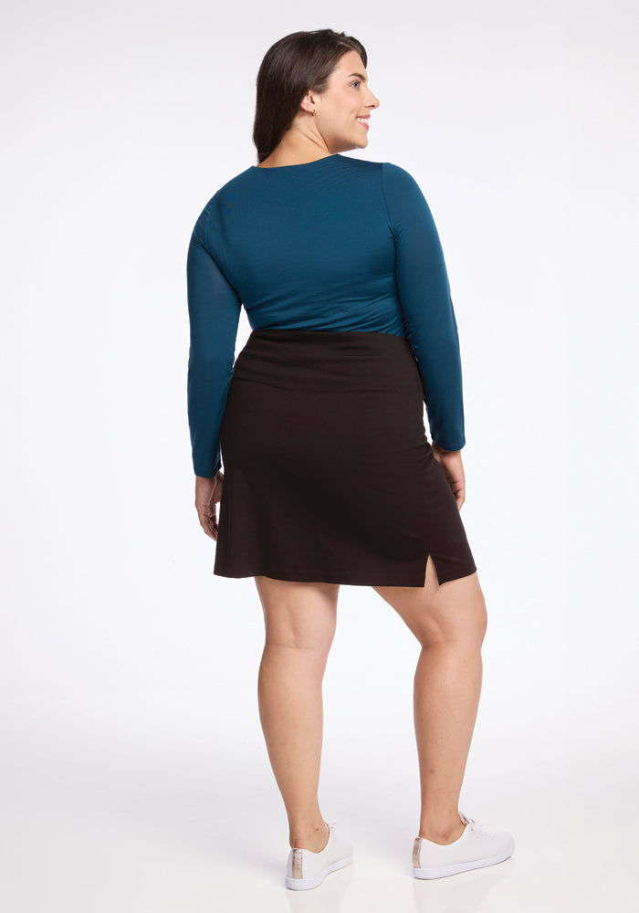 A woman stands facing away, clad in a teal long-sleeve top paired with the Woolx Monroe Knee Length Skirt in French Roast, featuring a side slit. Her long brown hair cascades down her back, and she completes the look with white sneakers. The moisture-wicking fabric ensures her comfort against the pristine white backdrop.