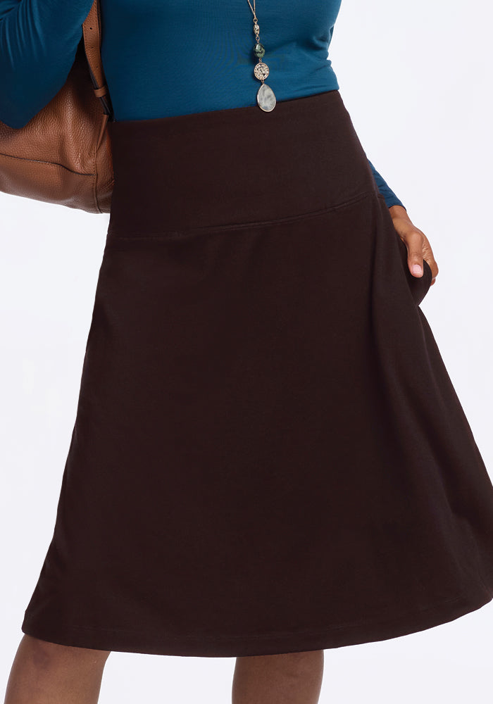 A person wearing a teal long-sleeve top with a necklace and the Marilyn Flowy Midi Skirt in French Roast by Woolx, holding a brown bag in one hand while the other rests casually in the skirt pocket.