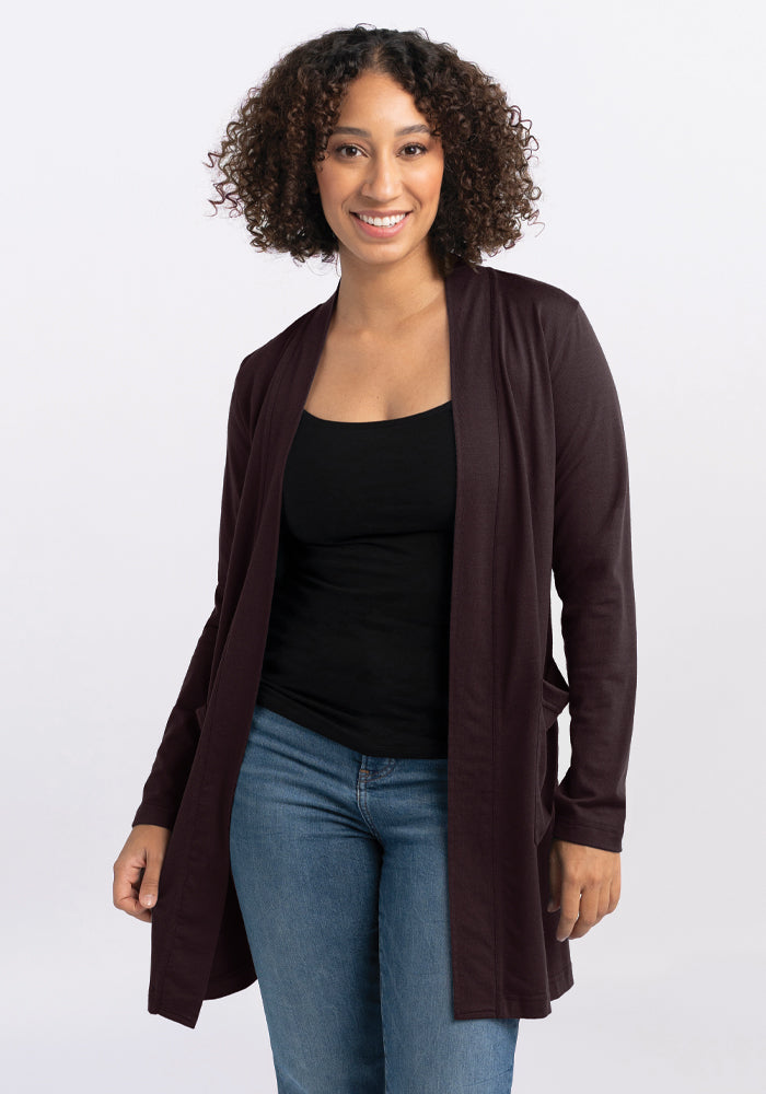 A person with curly hair smiles while wearing the Woolx Paisley Cardigan in French Roast, crafted from an Australian Merino Wool blend, over a black top and blue jeans, standing against a plain white background. 