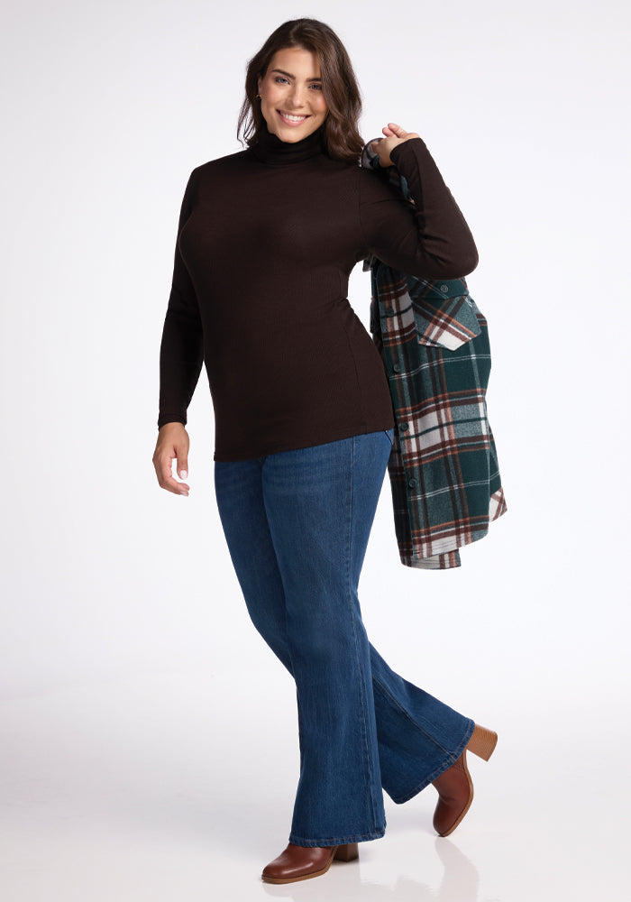 A person smiles while holding a green plaid jacket over their shoulder. They are wearing the Sage Ribbed Turtleneck in French Roast from Woolx, along with blue jeans and brown boots, standing against a plain white background.