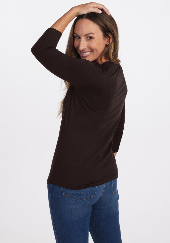 A person with long hair, wearing a Woolx Jenny 3/4 Sleeve Crew Neck in French Roast and blue jeans, smiles and looks over their shoulder against a plain white background.