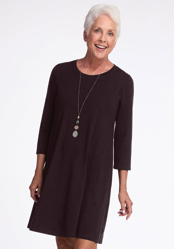 An older woman with short white hair stands gracefully, smiling in the Rissa A Line Dress in French Roast by Woolx, paired with a long necklace, set against a plain white background.