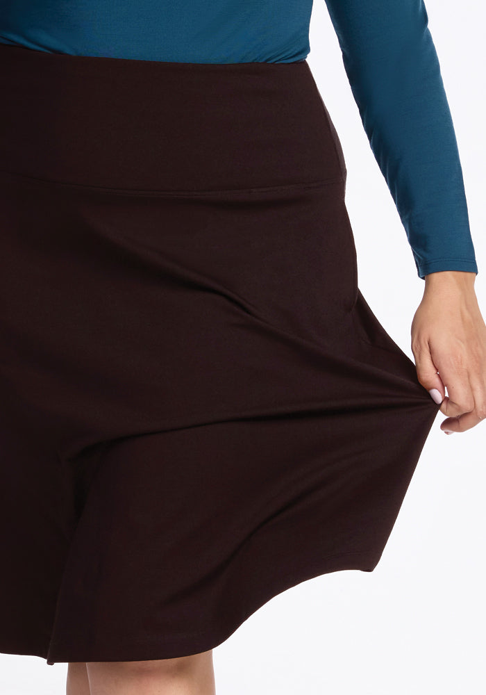 A person wearing a long-sleeved teal shirt gracefully tugs at the fabric of the Marilyn Flowy Midi Skirt - French Roast from Woolx, highlighting its stretchiness and elegant A-line silhouette against a plain white background.