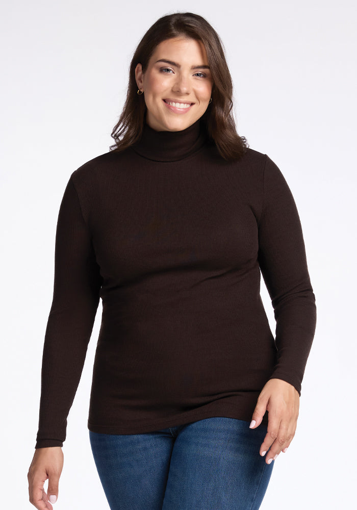 A person with long hair smiles while wearing the Sage Ribbed Turtleneck in French Roast from Woolx and blue jeans. The ribbed texture adds dimension, enhancing their casual look as they stand against a plain white background. 