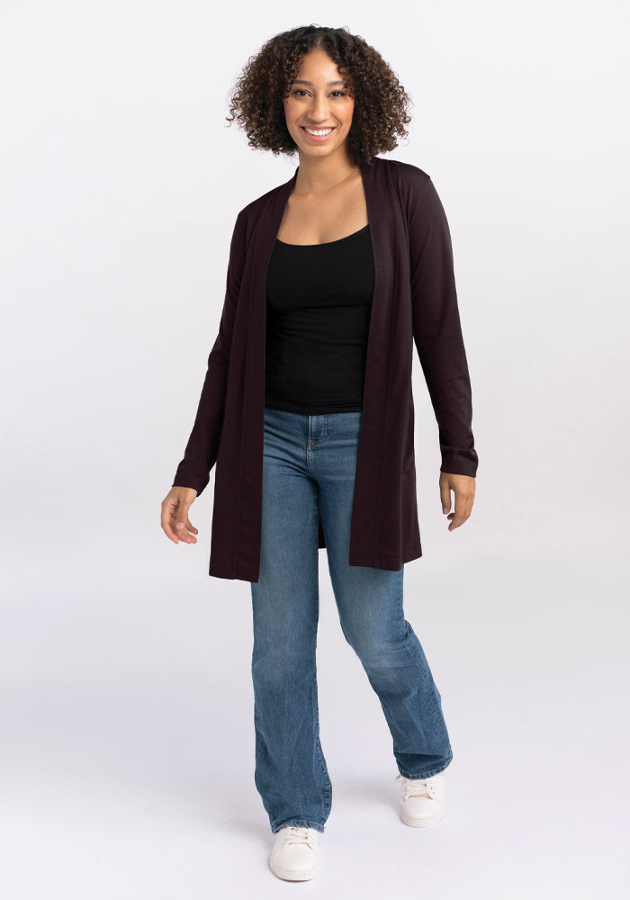 A person with curly hair is smiling and walking towards the camera, wearing a black top, a Woolx Paisley Cardigan in French Roast, blue jeans, and white shoes. The background is a plain, light color.