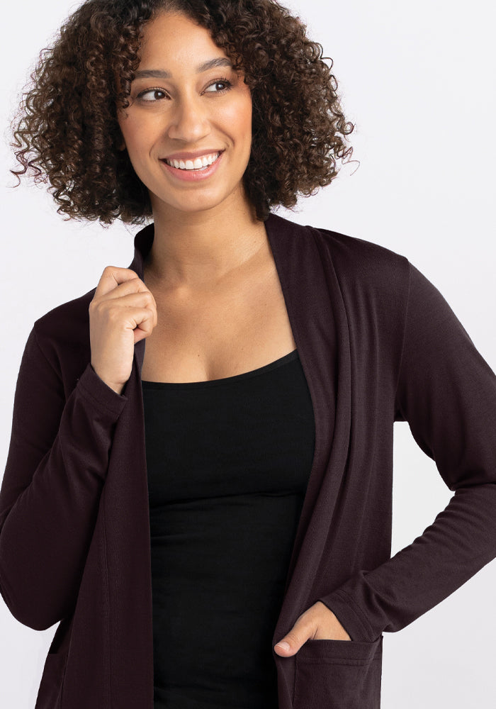 A person with curly hair smiles and looks to the side, dressed in a black top paired with the Paisley Cardigan - French Roast by Woolx. The background is plain white.