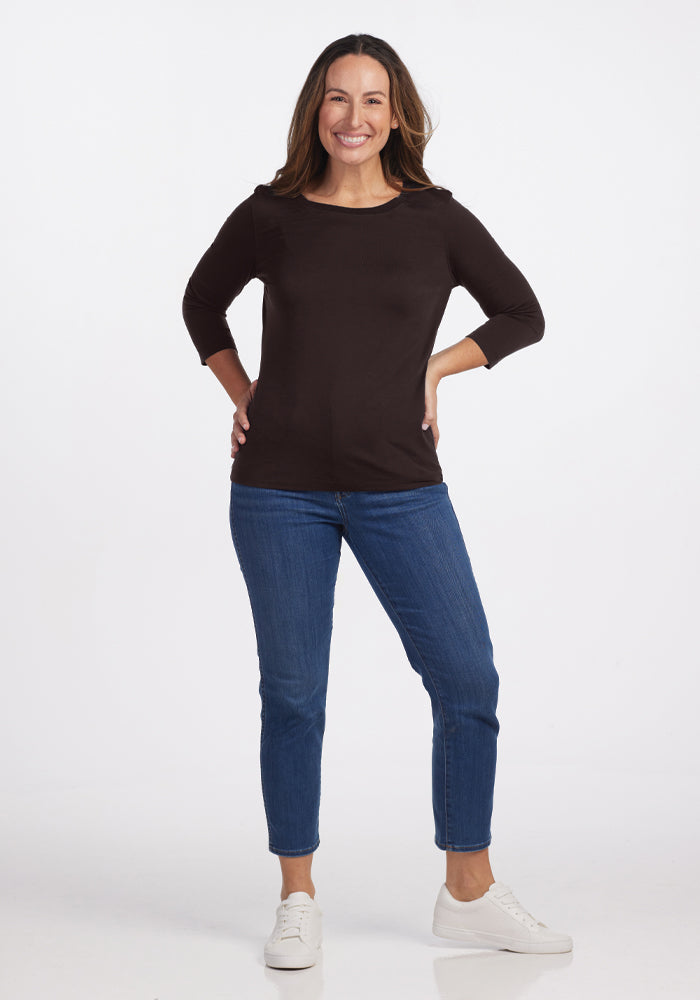 A person with long, dark hair stands smiling against a plain background. They are wearing a Jenny 3/4 Sleeve Crew Neck in French Roast by Woolx, blue jeans, and white sneakers, with their hands on their hips.