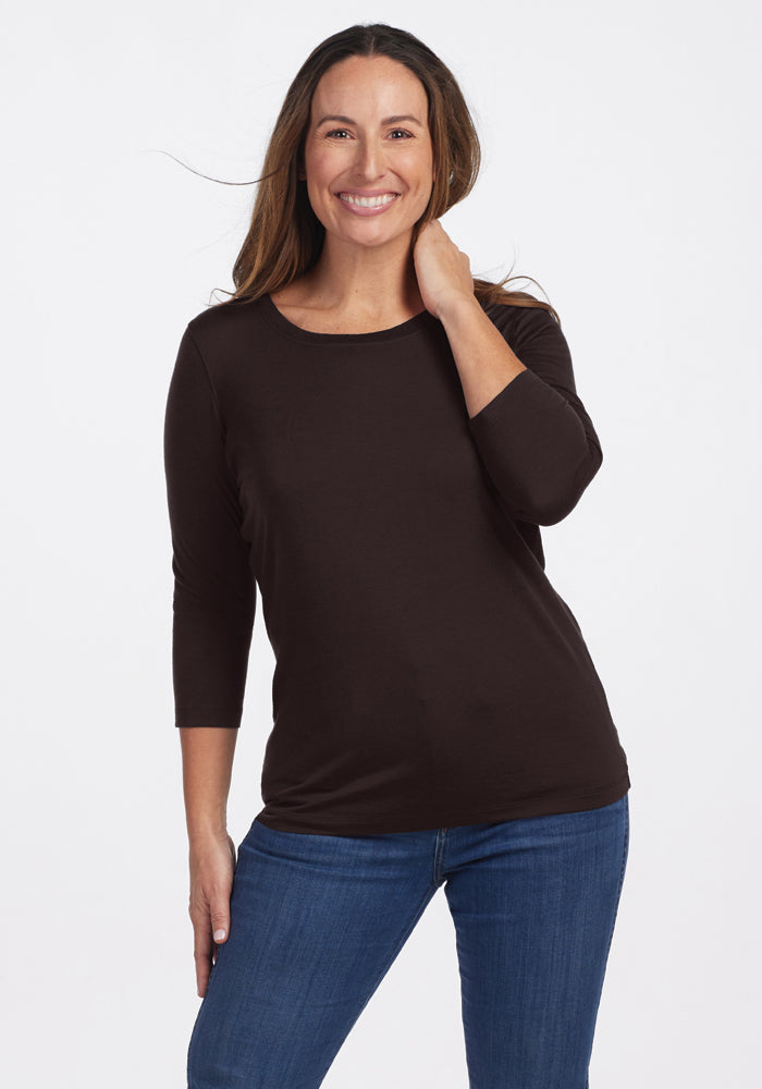 A person smiles while posing in a casual outfit, wearing the Woolx Jenny 3/4 Sleeve Crew Neck in French Roast and blue jeans. They have long hair and stand against a plain white background. 