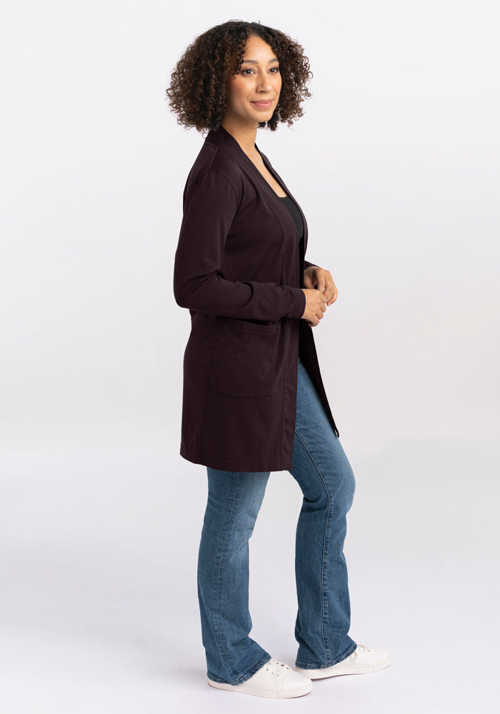 A person with curly hair is standing sideways, wearing a Woolx Paisley Cardigan in French Roast, paired with jeans and white shoes. They are posing confidently against a plain white background.