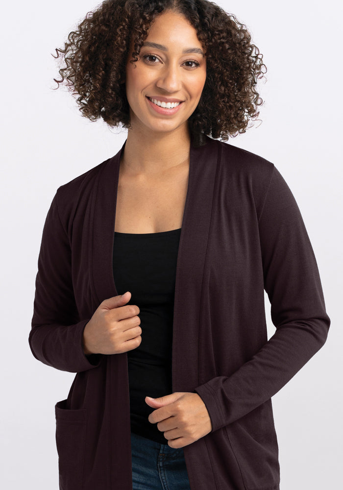 A person with curly hair smiles while wearing a Woolx Paisley Cardigan in French Roast over a black top. They stand against a plain white background, holding the edge of the cardigan with one hand.