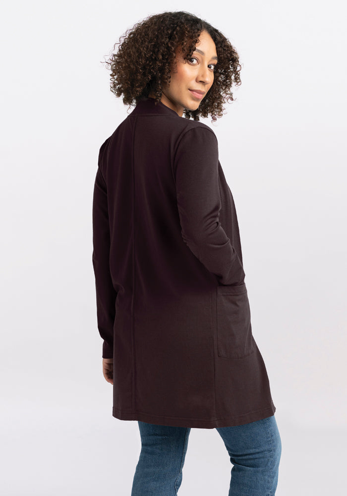 A woman with curly hair stands looking over her shoulder, wearing a long-sleeved Paisley Cardigan in French Roast from Woolx. The cardigan is crafted from Australian Merino Wool, and she pairs it with blue jeans, her hands tucked into the cardigan pockets. The backdrop is a plain, light gray.