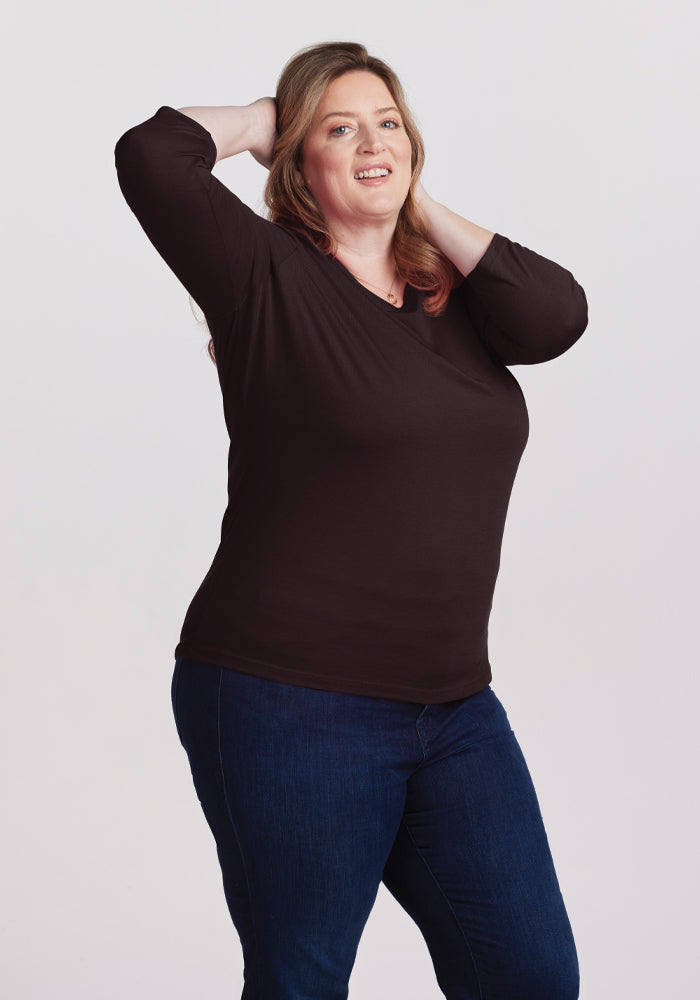 A person with long hair is posing confidently with both hands behind their head, adorned in the chic Jenny 3/4 Sleeve Crew Neck in French Roast by Woolx, and dark blue jeans. The background's plain light color accentuates the elegance of lightweight Australian Merino Wool, enhancing this stylish ensemble.