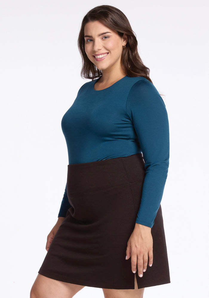 A woman with long brown hair is smiling and wearing a teal long-sleeve top and the Woolx Monroe Knee Length Skirt in French Roast. The knee-length design of the dark brown skirt looks stylish as she stands against a plain white background.