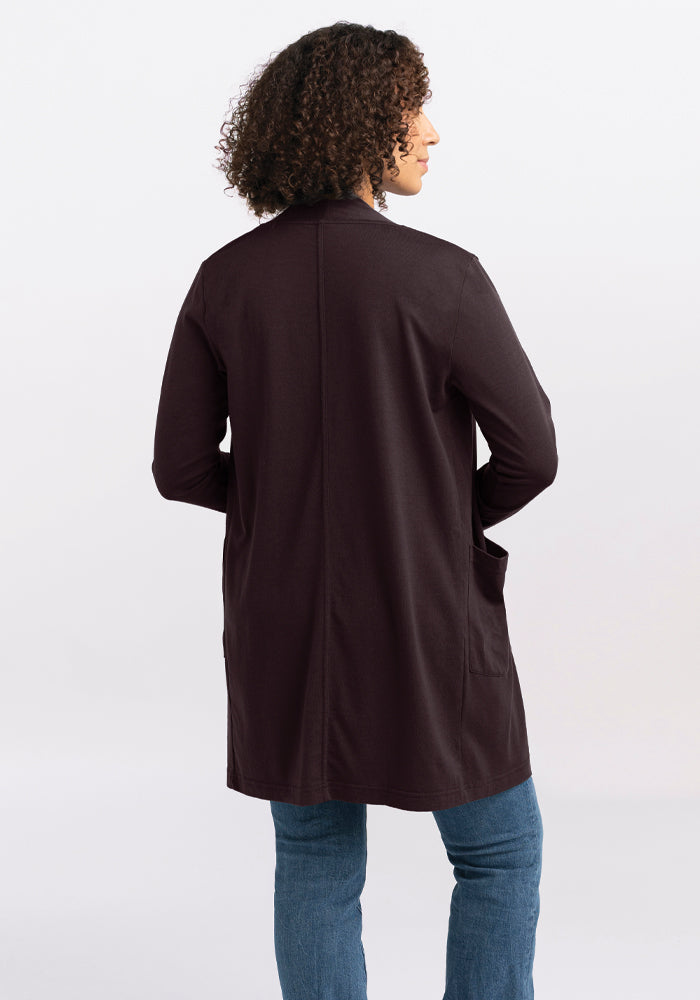 A person with curly hair is standing with their back to the camera, wearing a long, dark Paisley Cardigan - French Roast made of Australian Merino Wool from Woolx, paired with blue jeans against a plain background.