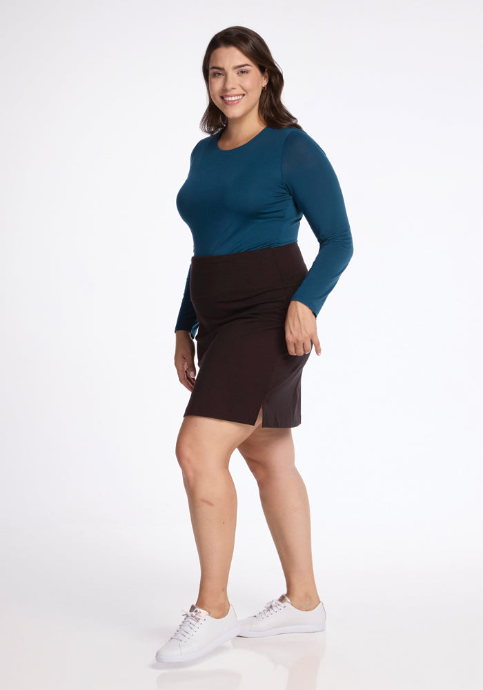 A woman stands smiling, dressed in a teal long-sleeve top and the Monroe Knee Length Skirt in French Roast by Woolx. Her ensemble is complemented by white sneakers, perfectly balancing style and comfort against a plain white background that highlights the chic look.