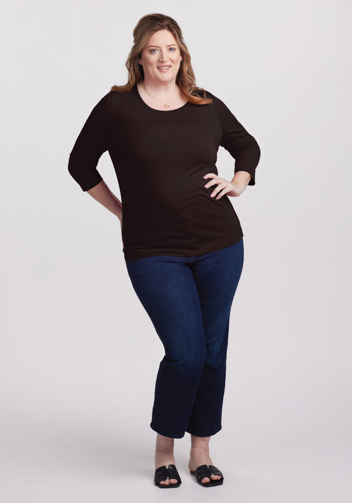 A person with long hair, wearing a Woolx Jenny 3/4 Sleeve Crew Neck in French Roast and dark blue jeans, stands with one hand on their hip. They are also wearing black sandals against a plain white background.