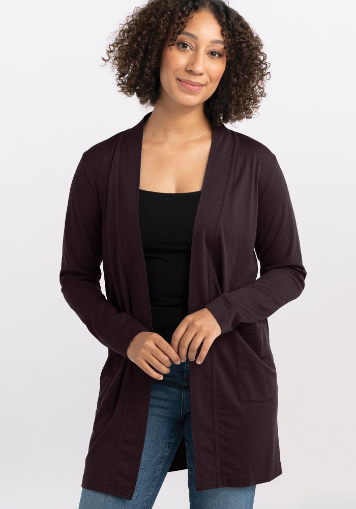 A person with curly hair is wearing the Woolx Paisley Cardigan in French Roast over a black top and blue jeans. They are standing against a plain background, smiling gently at the camera with hands clasped in front.