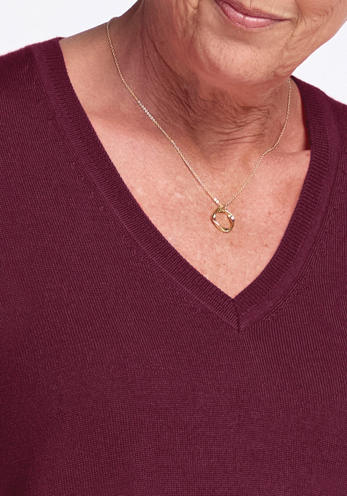 A person is wearing the Francesca Sweater in Cabernet Crush by Woolx, a luxurious Merino wool V-neck sweater, paired with a gold necklace featuring a simple loop pendant. The image highlights the intricate details of both the clothing and accessory against a neutral background.