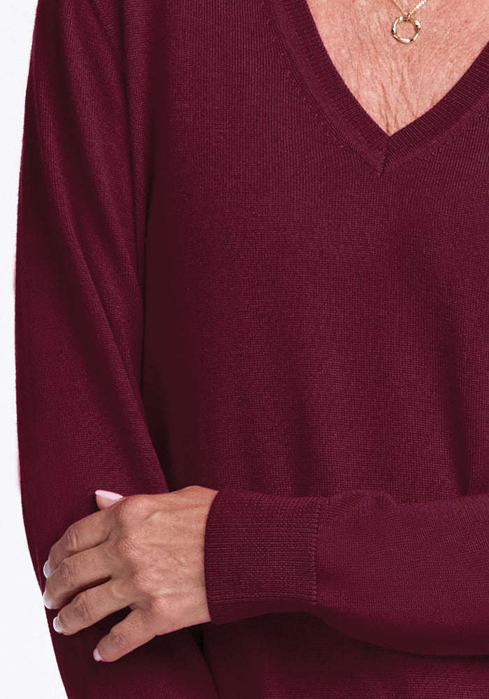 A person in the Francesca Sweater - Cabernet Crush by Woolx, made from soft merino wool, is shown with their arms crossed. They wear a gold necklace featuring a simple circular pendant. The image highlights the upper body, emphasizing the sweater's texture and luxurious material.