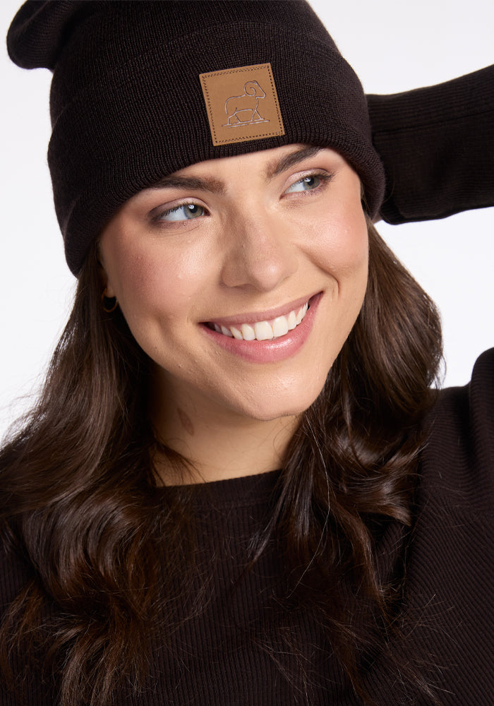 A woman with long brown hair smiles while wearing the Woolx Baylor Beanie in French Roast, distinguished by its brown patch. Her black sweater complements the stretchy foldable ear cuff as she poses with one hand adjusting her hat against a plain white background.