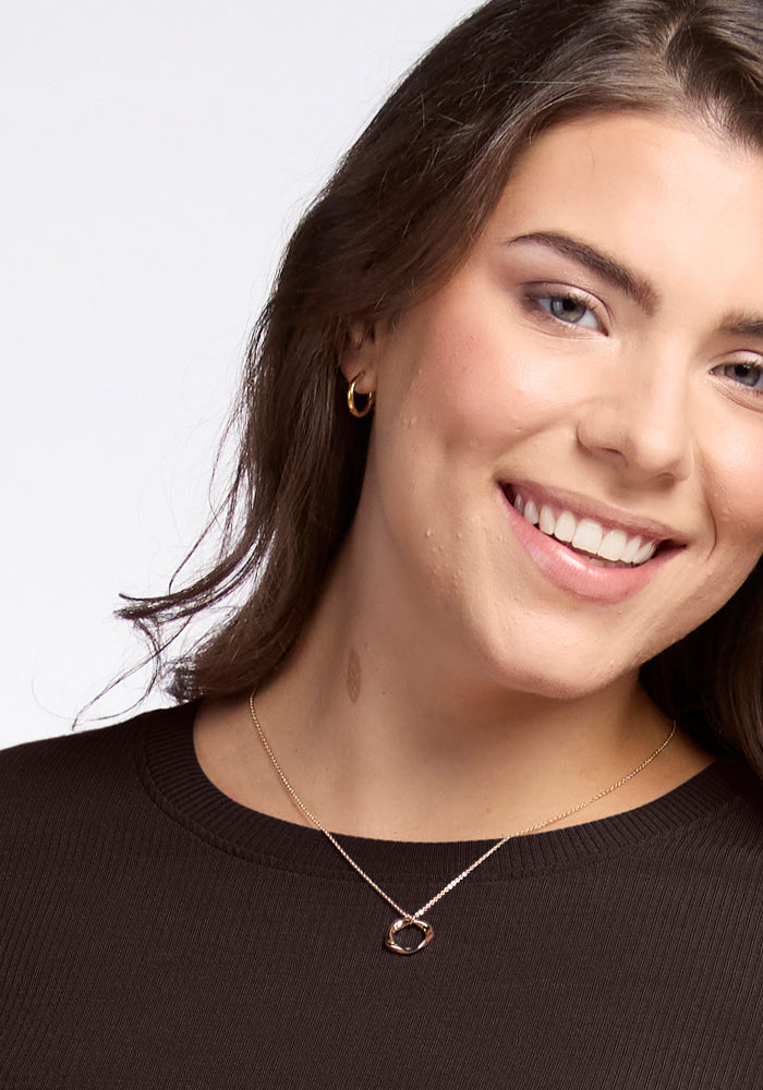 A woman with long brown hair wearing a French Roast-colored Hadley Ribbed Crew by Woolx and a gold necklace smiles at the camera. She accessorizes with gold hoop earrings and has opted for a light makeup look. The plain, light background accentuates her stylish apparel made from luxurious Australian Merino Wool.