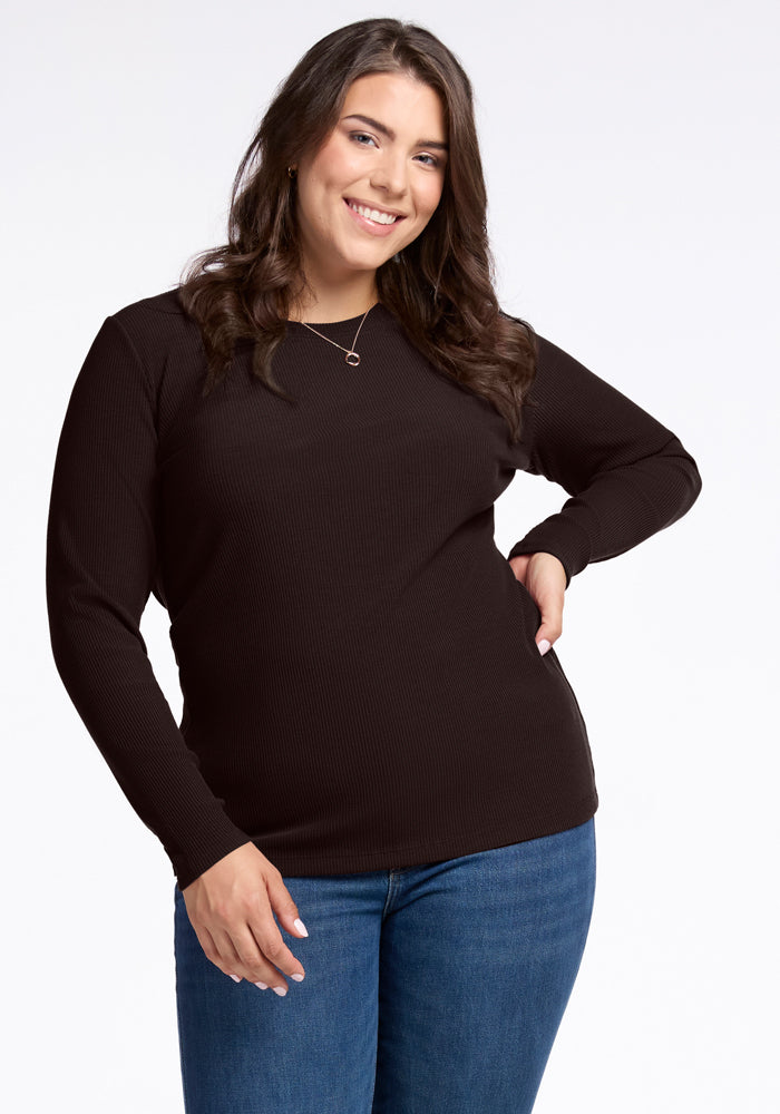 A person with long hair is smiling brightly, wearing a French Roast Hadley Ribbed Crew top by Woolx and blue jeans, standing against a plain white background. They have one hand on their hip and are adorned with a stylish necklace. 