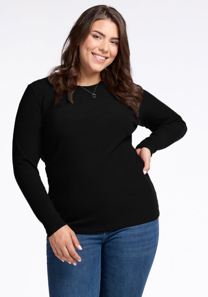 A person with long brown hair smiles at the camera, wearing the Woolx Hadley Ribbed Crew in black and blue jeans. They have one hand on their hip and the other arm relaxed by their side, exuding a luxurious feel against the plain white background. 