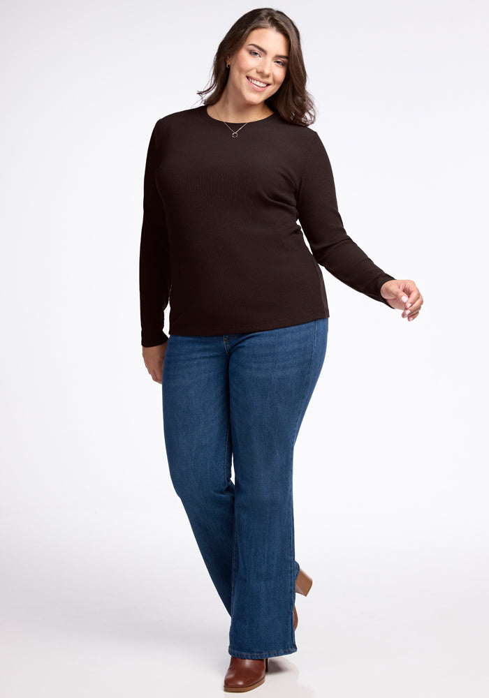 A woman is posing in a French Roast Hadley Ribbed Crew top by Woolx and blue jeans. She is smiling and standing against a plain white background, with her left arm slightly bent and her hand resting near her hip. Her look is completed with brown boots.