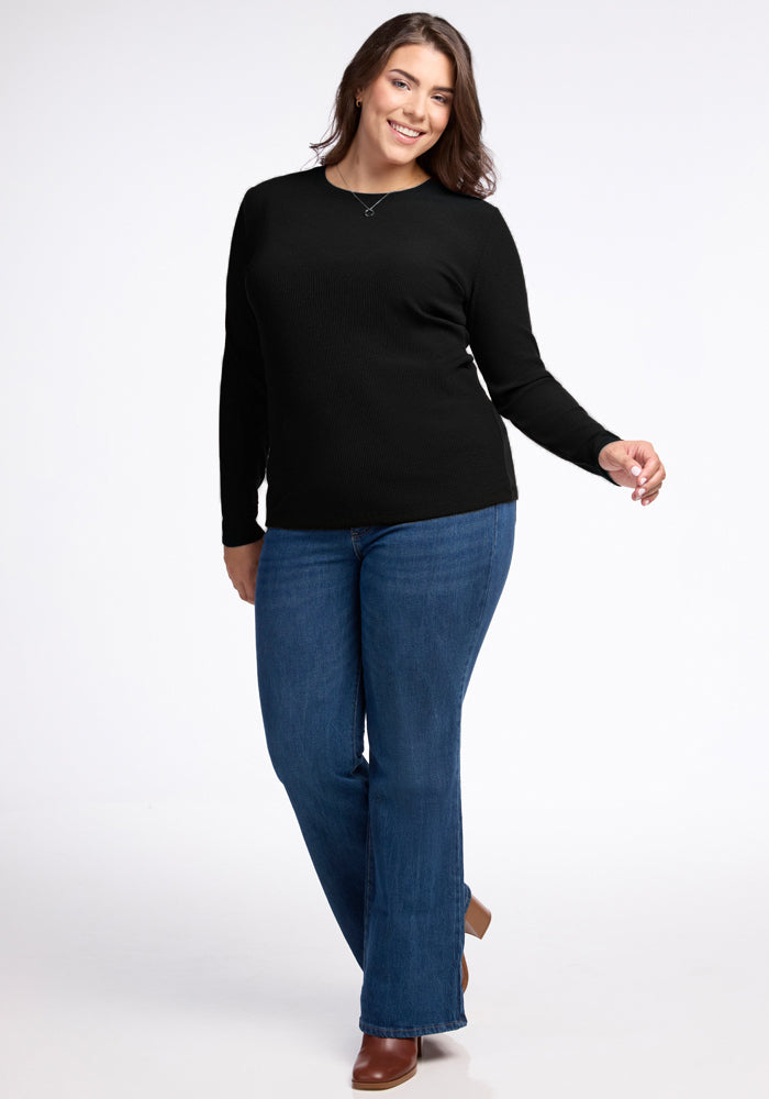 A woman wearing the Woolx Hadley Ribbed Crew in black and blue jeans stands smiling. She confidently poses with one hand on her hip and the other by her side, complemented by brown shoes. The plain white background highlights the luxurious feel of her outfit.