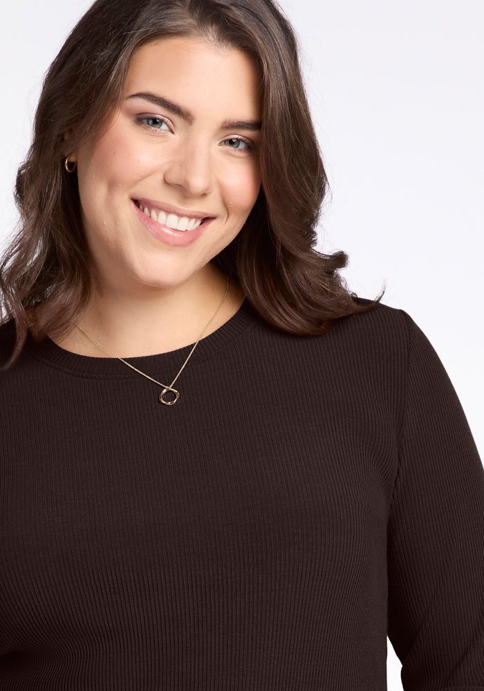 A woman with long brown hair smiles warmly at the camera. She is wearing the Woolx Hadley Ribbed Crew in French Roast, crafted from Australian Merino Wool, featuring a stylish crew neckline, complemented by a necklace with a round pendant against a light background.