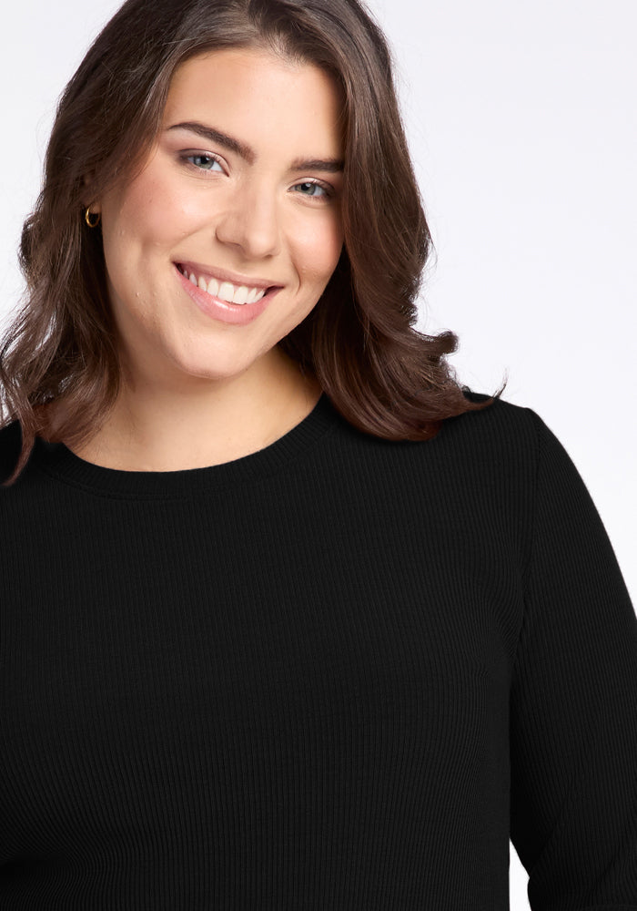 A person with shoulder-length brown hair smiles warmly at the camera. They are wearing the Hadley Ribbed Crew in black from Woolx, featuring a simple long-sleeved design that adds a touch of elegance with its textured fabric. The background is plain and white, creating a bright and clear setting.