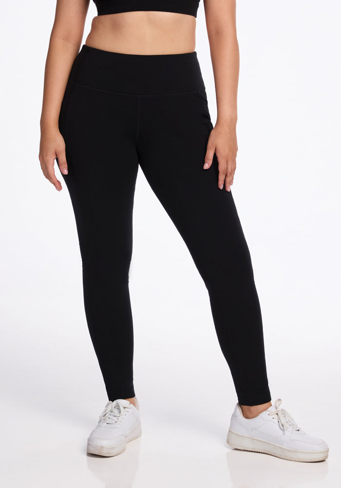 A person stands against a white background, wearing a black crop top, Woolx Frost Duralite® Leggings in black, and white sneakers. The image focuses on their midsection and legs, showcasing a casual and sporty outfit. 