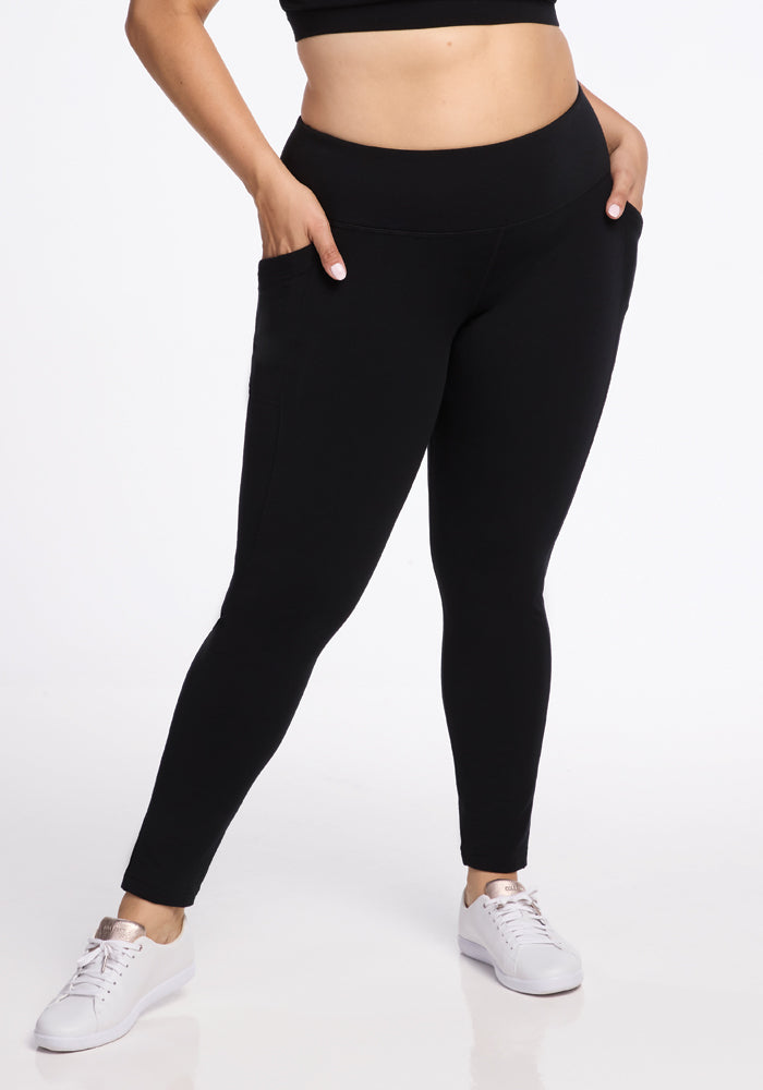 A person is wearing Woolx Frost Duralite® Leggings in black, with their hands in the side pockets. They are also sporting a black crop top and white sneakers. These warm leggings function as the perfect baselayer for any outfit. The background is plain white. 