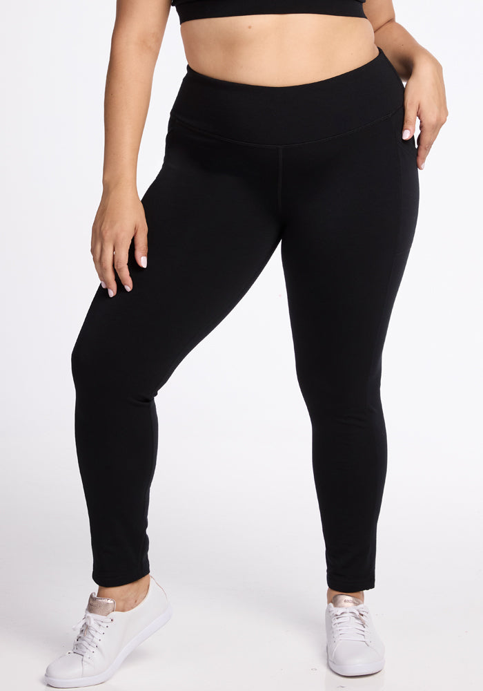 A person with light skin is wearing Woolx's Frost Duralite® Leggings in black paired with a black crop top. They are also wearing white sneakers against a plain white backdrop. The individual stands with one arm hanging loose while the other rests on their hip. 