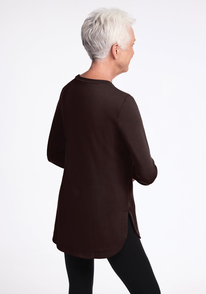 A person with short white hair, wearing the Woolx Blair Tunic in French Roast and black pants, is facing away from the camera on a plain white background. This odor-resistant outfit makes an ideal travel companion.