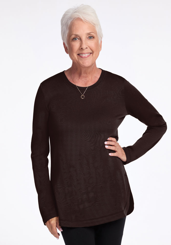 An older adult with short gray hair is smiling while wearing the Woolx Blair Tunic in French Roast and a necklace. The odor-resistant Merino wool drapes comfortably as they stand with one hand on their hip against a plain white background. 