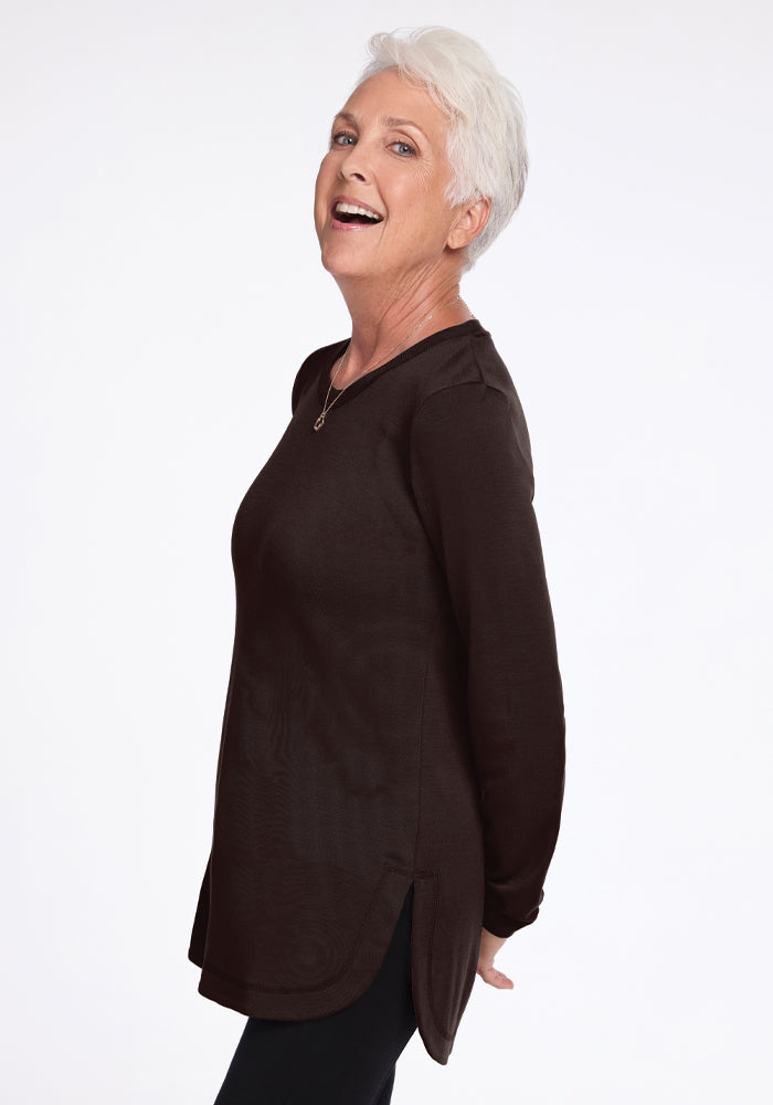 A smiling older woman with short white hair stands in profile against a plain white background. She is wearing the odor-resistant Blair Tunic in French Roast from Woolx and black pants, with her hands clasped behind her back.