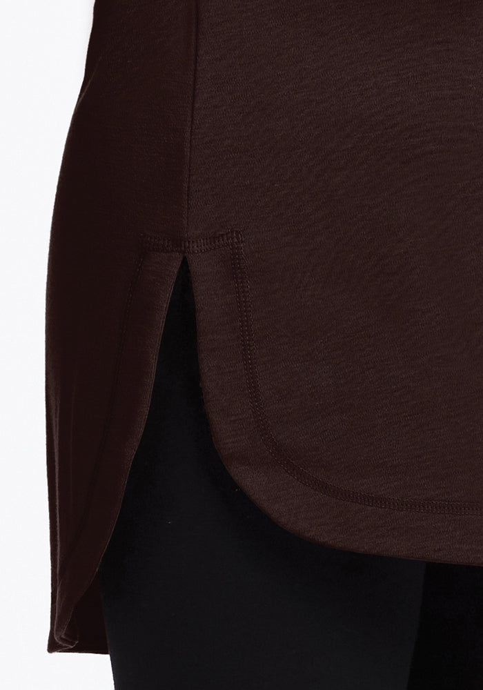 Close-up of the Woolx Blair Tunic in French Roast, a lightweight travel piece with a curved hem and side slit, elegantly draped over black leggings. Made from odor-resistant merino wool, it offers a soft texture with visible stitching details along the edge.