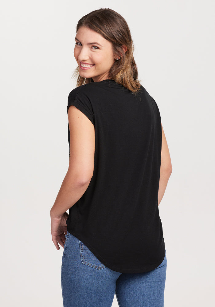 A person with shoulder-length hair smiles while looking over their shoulder, dressed in the Woolx Gabi Tunic Top in black paired with blue jeans, set against a plain white background.