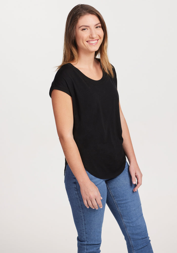 A person with long hair smiles while standing, wearing the versatile Gabi Tunic Top in black by Woolx and blue jeans against a plain white background.