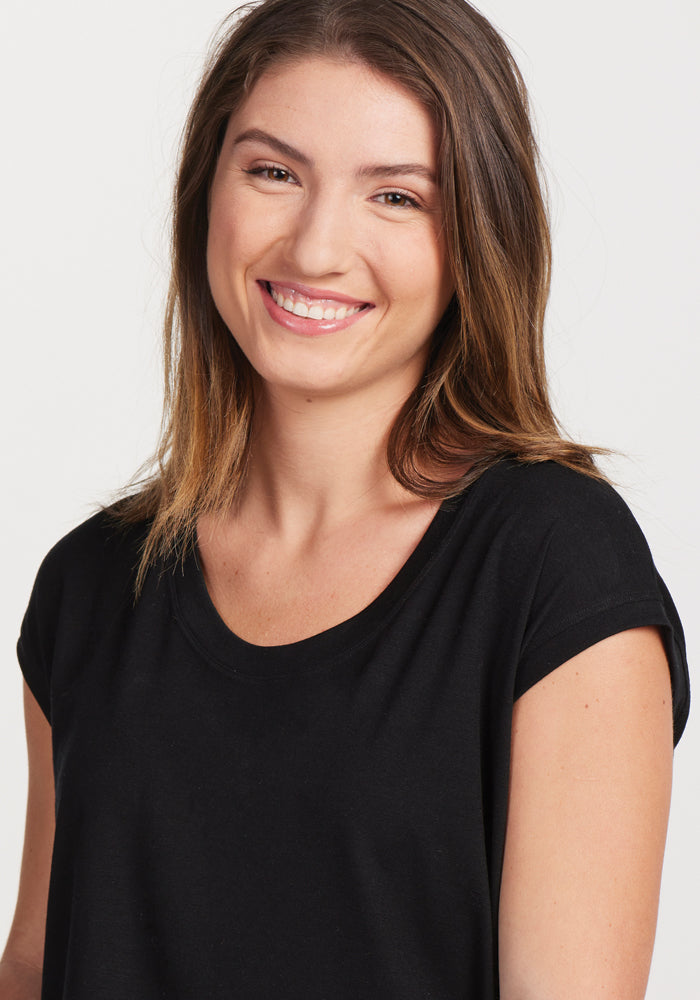 A smiling person with long brown hair stands against a plain background, wearing the Gabi Tunic Top in black by Woolx, exuding effortless style.