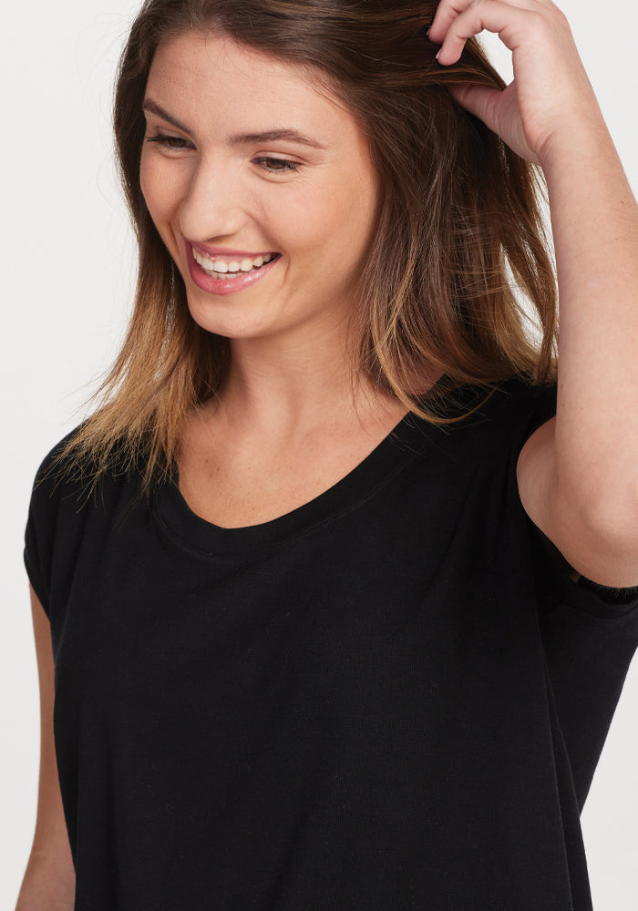 A woman with long brown hair is smiling as she touches her hair, wearing the Gabi Tunic Top in black by Woolx. Her cheerful expression stands out against the plain white background.