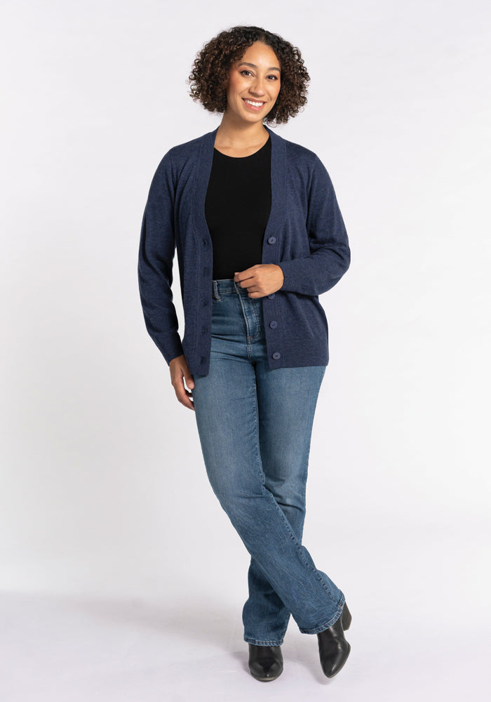 A person wearing a black shirt, the Gia Sweater in Twilight Storm by Woolx, and blue jeans stands smiling against a plain background. They have curly hair and are wearing black shoes.